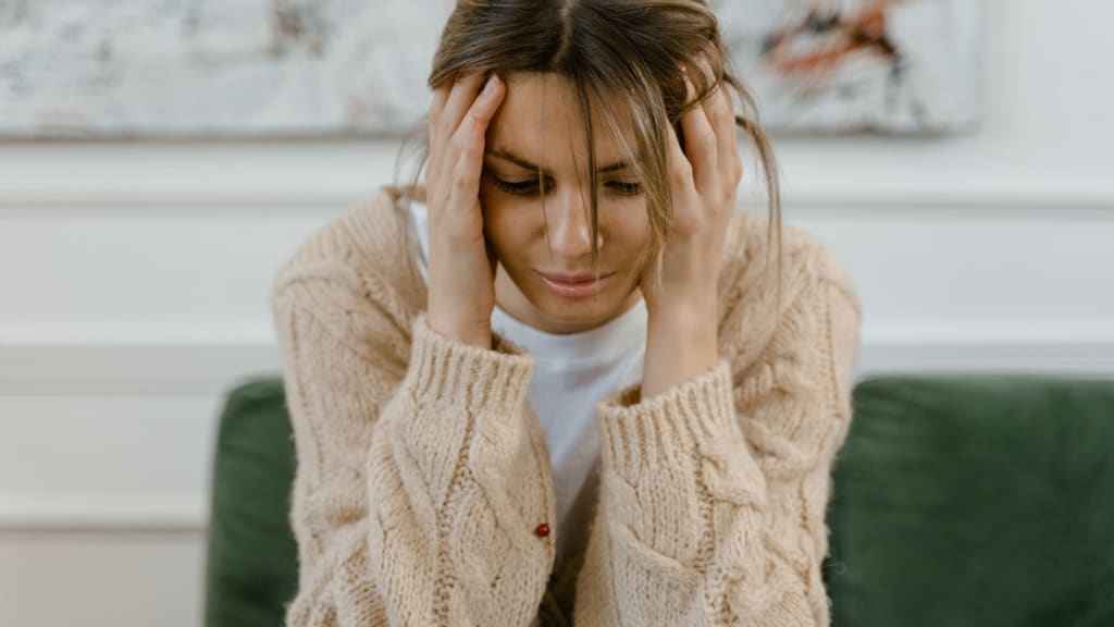 A woman sits on a green couch, holding her head in her hands and looking down, appearing stressed or upset as if she might benefit from speaking with a therapist.